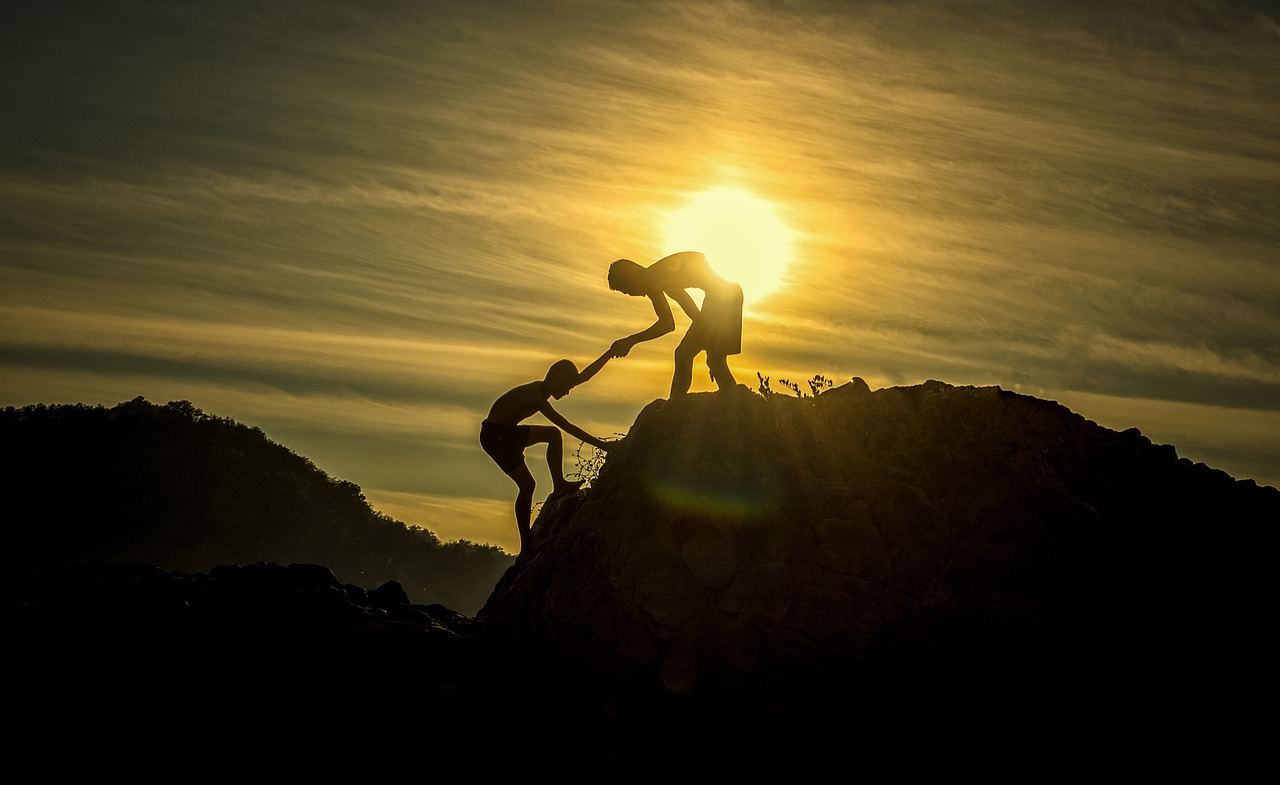 Una persona ajuda a una altra a coronar un cim sota el sol que comença a sortir